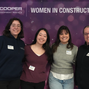 Four attendees at the Cooper Women in Construction breakfast smile in front of a purple photo wall.
